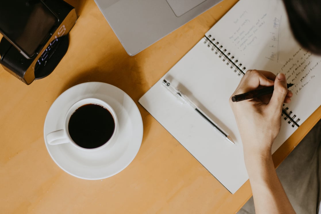 Student Writing on Notebook Beside Coffee Cup